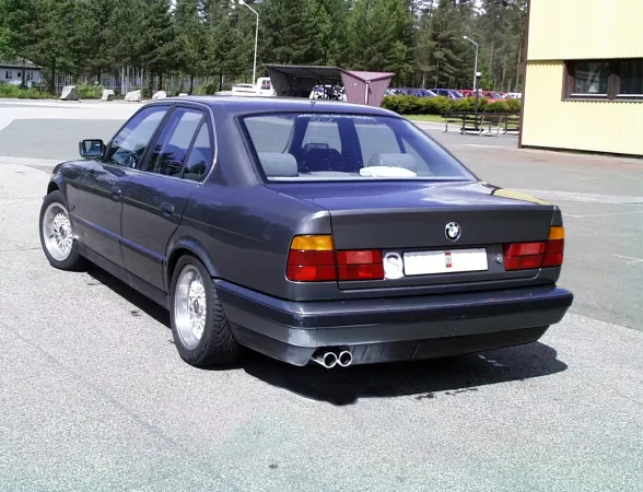 Vue arrière d'une BMW E34 gris foncé. On voit le système d'échappement sport duplex en acier chromé avec 2x70mm sorties, installé sous le pare-chocs. La voiture est garée sur un parking, avec des arbres et un bâtiment en arrière-plan.